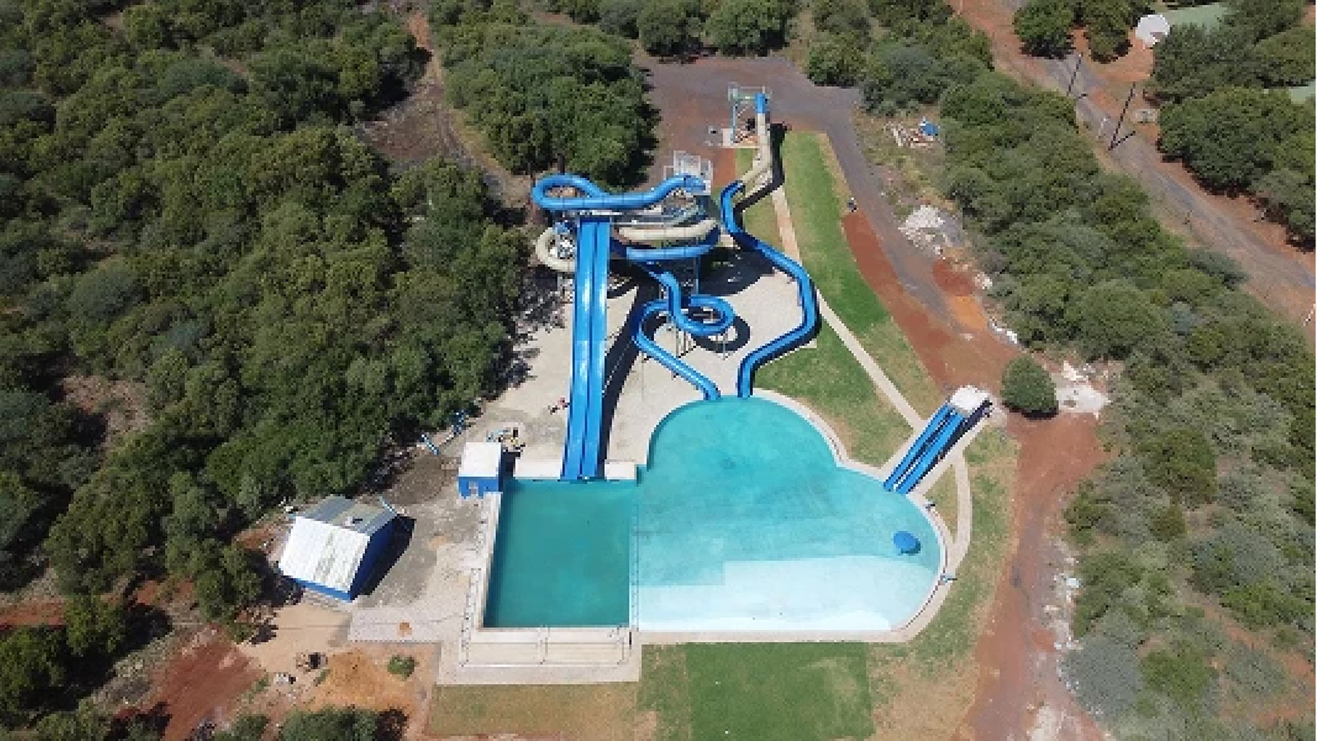 Aerial view of a water park featuring several tall, intertwining blue water slides leading into a large, clear pool, surrounded by lush greenery and paths.