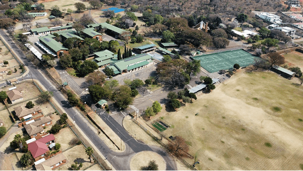 Aerial view of a school campus with multiple green-roofed buildings, tennis courts, and surrounding greenery. Roads border the campus, with houses nearby.
