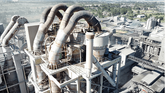 Industrial plant with large, rusted pipes and storage tanks, surrounded by machinery and greenery in the background. Aerial view shows scale.
