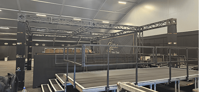 A metal stage with safety rails is set up in a large indoor venue. A black backdrop displays "Poker Tour" branding. The atmosphere is professional.